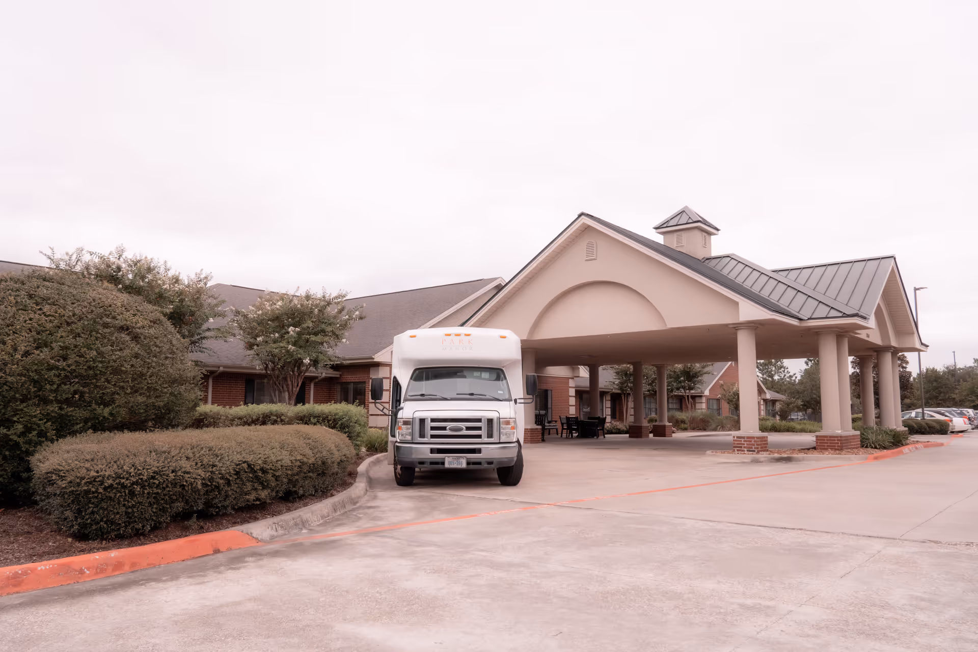 Covered entrance (porte-cochere) of a senior living facility with a white shuttle van parked in front and surrounding landscaping.