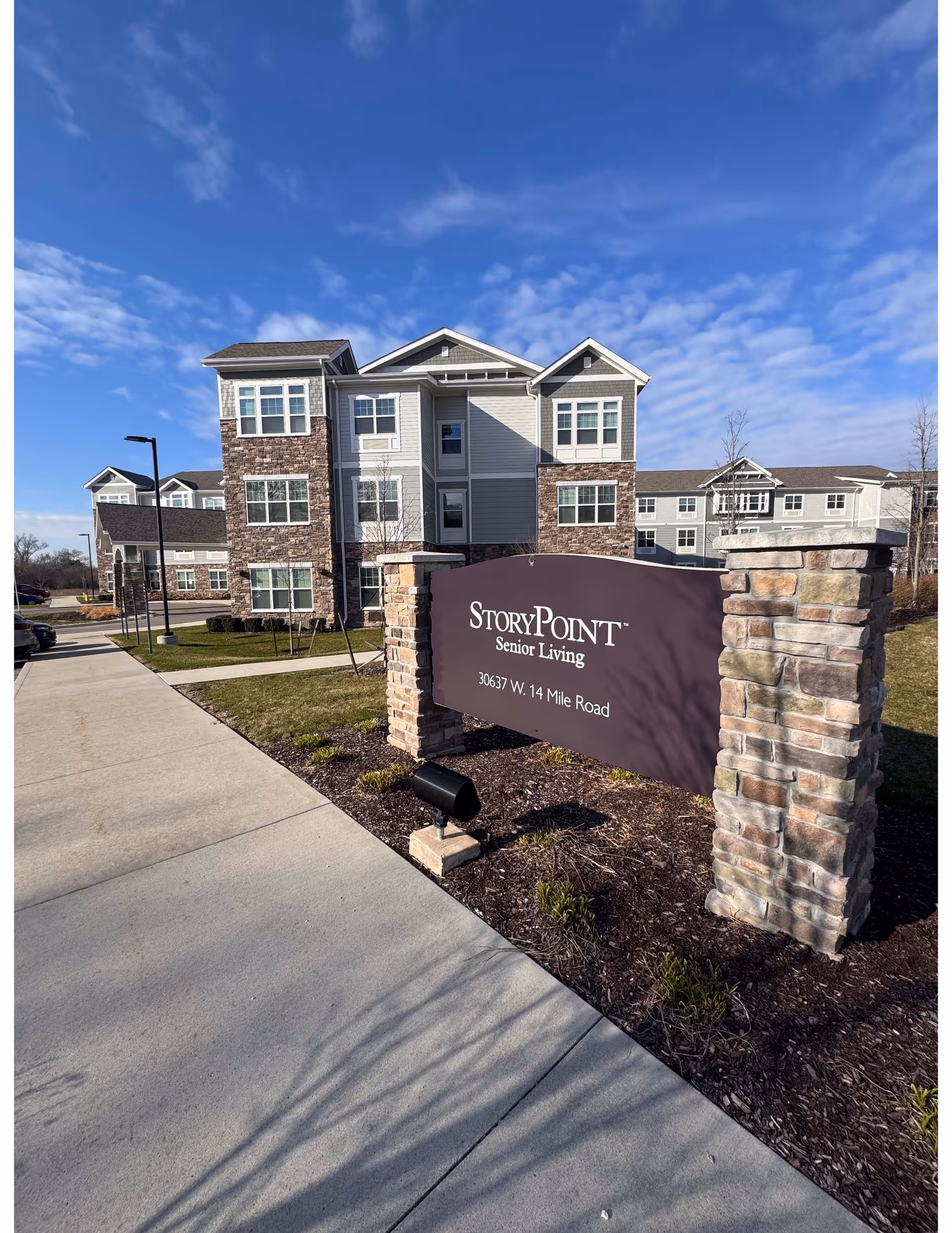 Exterior view of StoryPoint Senior Living facility in Farmington Hills, showing a multi-story building with stone and siding facade under a blue sky. In the foreground, there is a large sign with the facility name and address, surrounded by landscaping and a sidewalk.