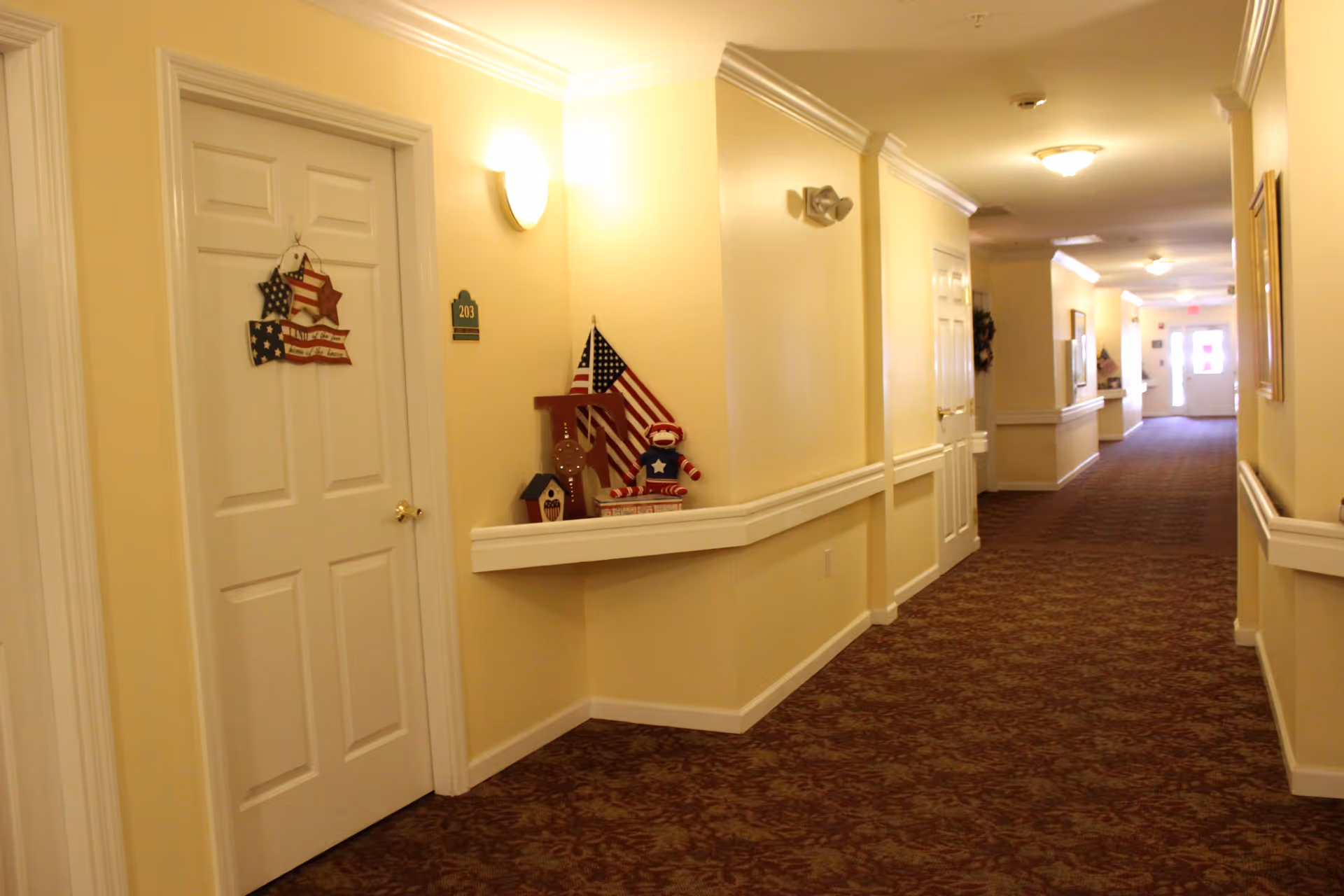 A well-lit hallway in an assisted living facility with cream-colored walls and patterned carpet. There are white doors along the hallway, one marked with the number 203 and decorated with patriotic items including an American flag and a star-shaped wall hanging. The hallway has handrails on both sides and light fixtures on the walls and ceiling.
