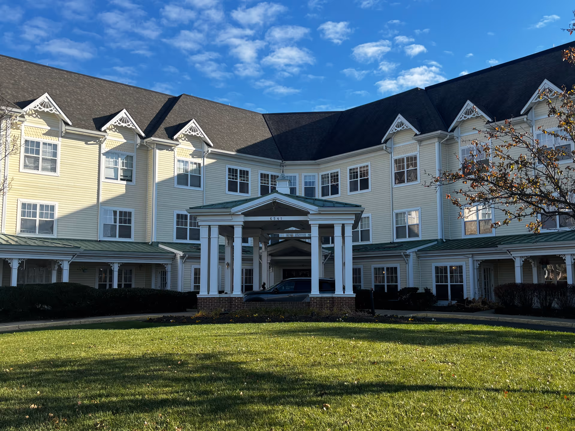Front exterior view of a large, three-story senior living facility building with cream-colored siding and a dark shingled roof under a blue sky with scattered clouds. The entrance features a covered portico with white columns and a green roof. There is a well-maintained lawn in the foreground and some bushes and trees around the building.