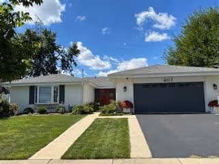 Single-story residential building with a white exterior, dark garage door, and a red front door. The house is surrounded by a well-maintained lawn and some shrubs, under a blue sky with scattered clouds.