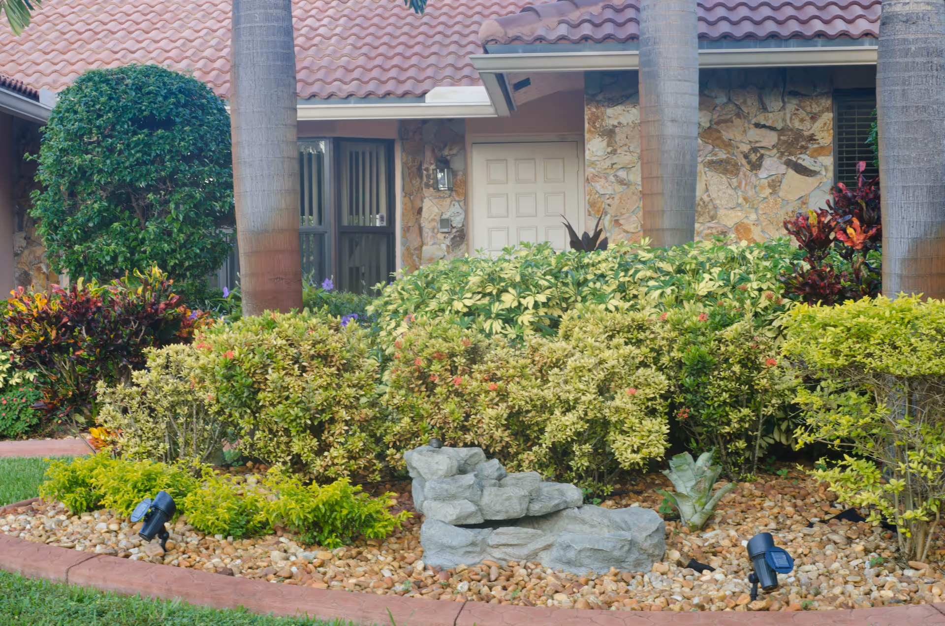 Front view of a residential building with a tiled roof, stone facade, and a white door partially obscured by lush green bushes and palm trees. The garden area in front features various plants, shrubs, decorative rocks, and two small black garden lights.