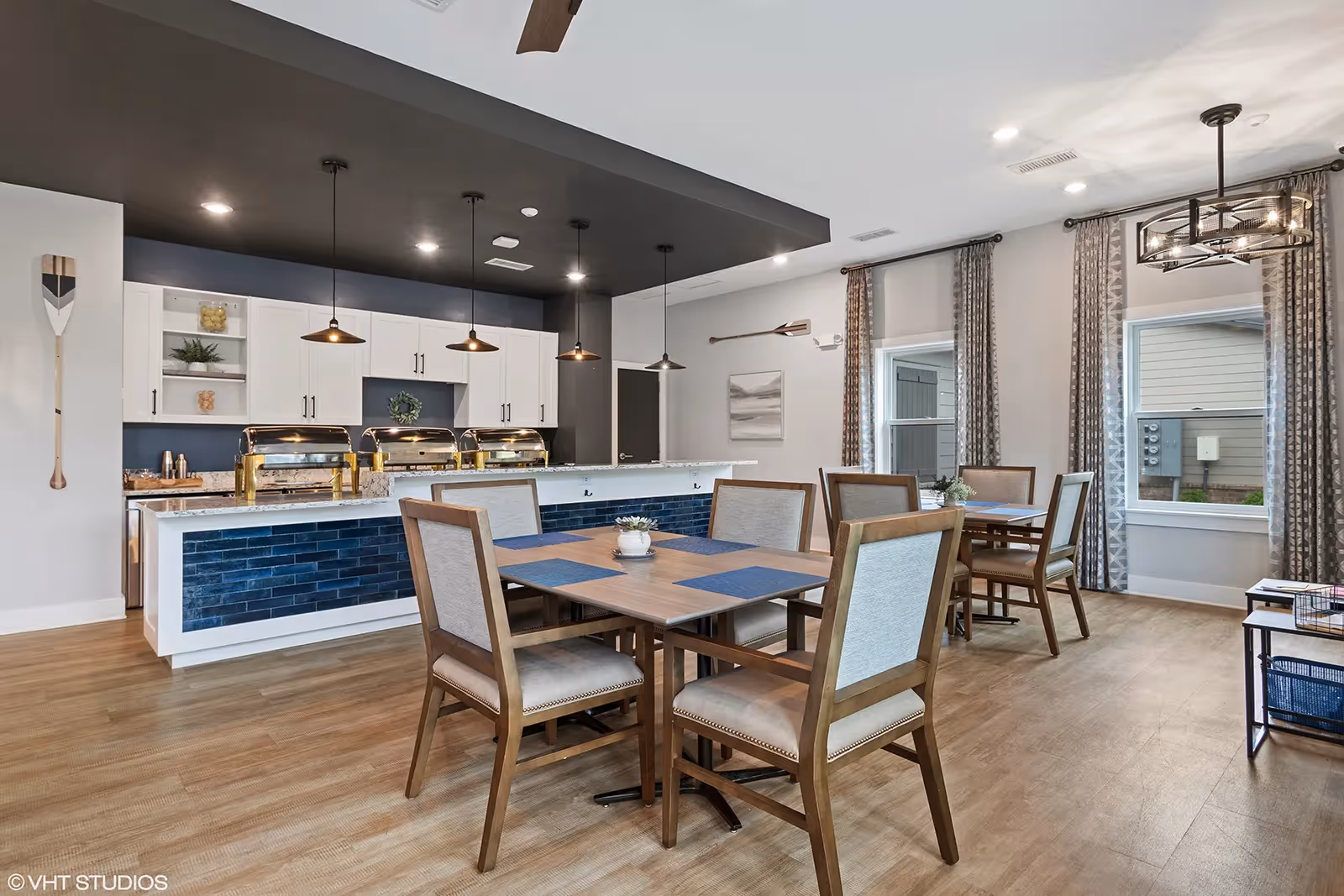 A modern dining area with wooden tables and cushioned chairs, featuring a buffet counter with chafing dishes and pendant lights above. The room has large windows with patterned curtains, light wood flooring, and decorative oars on the walls.