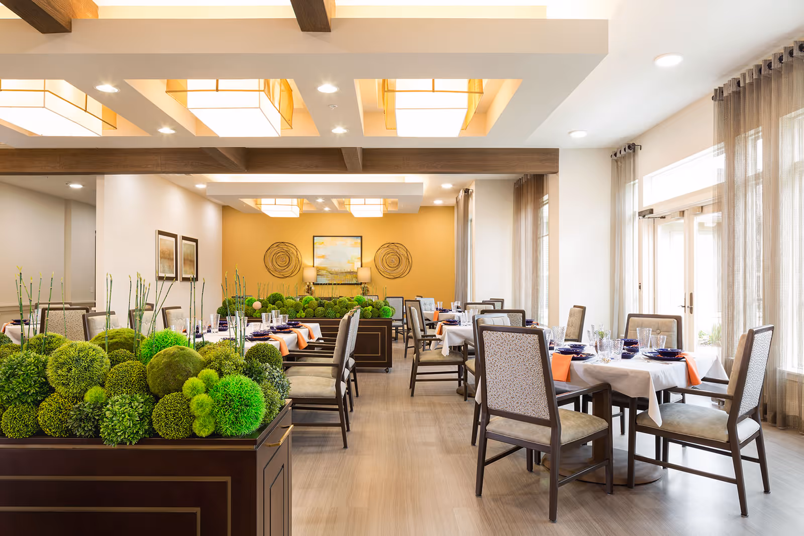 A bright and elegant dining room with several tables set with white tablecloths, orange napkins, blue plates, and clear glassware. The room features large windows with sheer curtains, modern ceiling lights, and decorative green plants arranged in planters dividing the space.