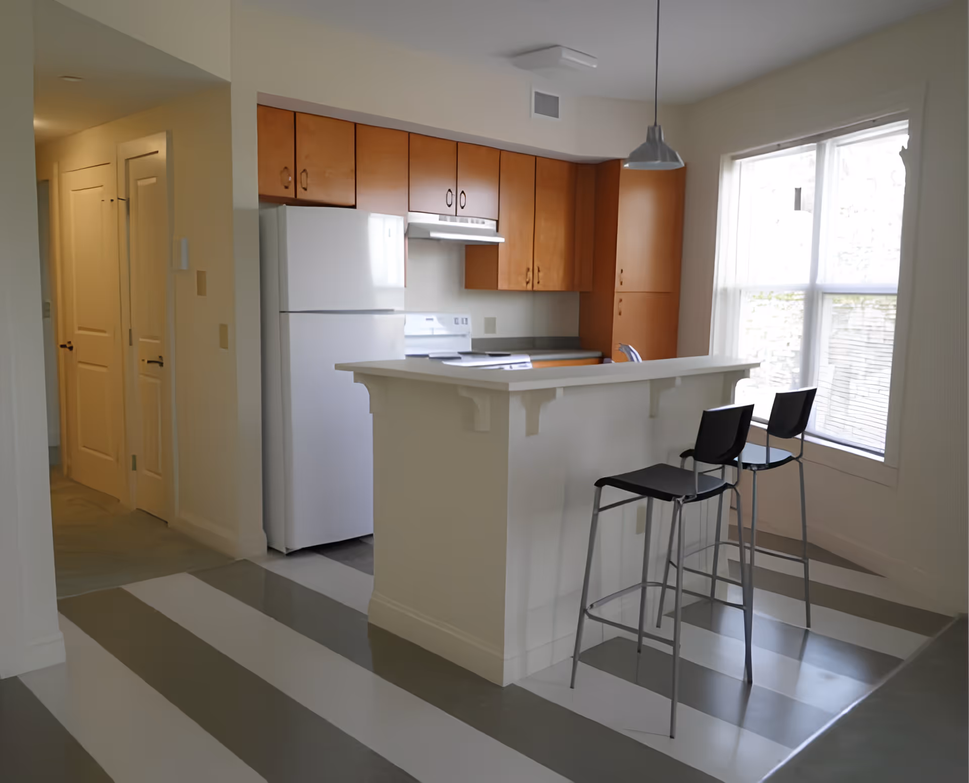 Bright kitchen with a breakfast bar, two tall stools, white appliances and wood cabinets next to a large window.