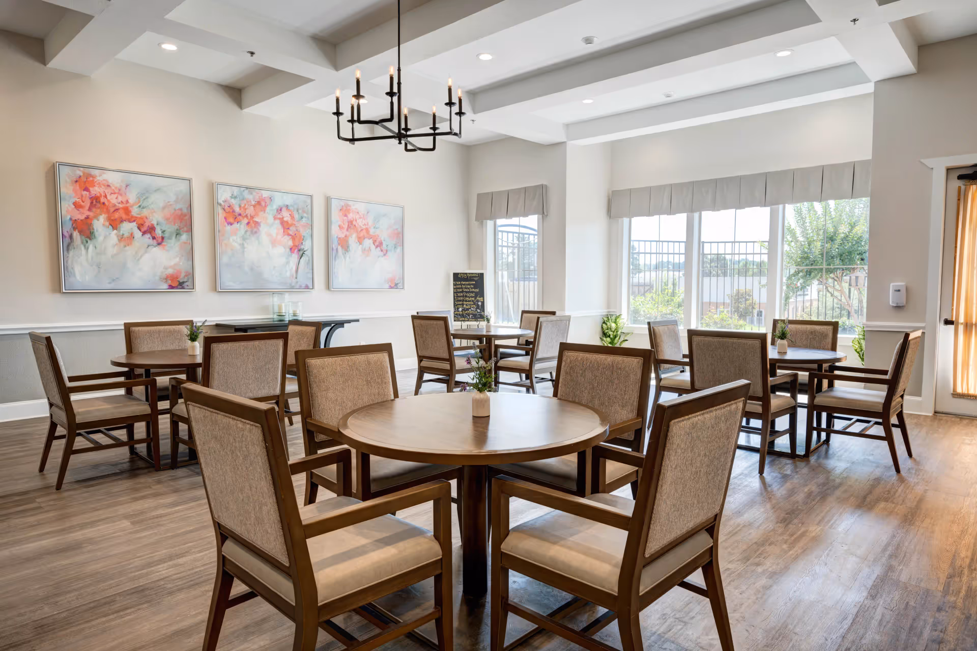 A bright and spacious dining room with several round wooden tables surrounded by cushioned chairs. The room features large windows with beige valances allowing natural light to fill the space. On one wall, there are three colorful abstract floral paintings, and a black chandelier hangs from the ceiling. The floor is covered with wood-style flooring, and small plants are placed on the tables and near the windows.