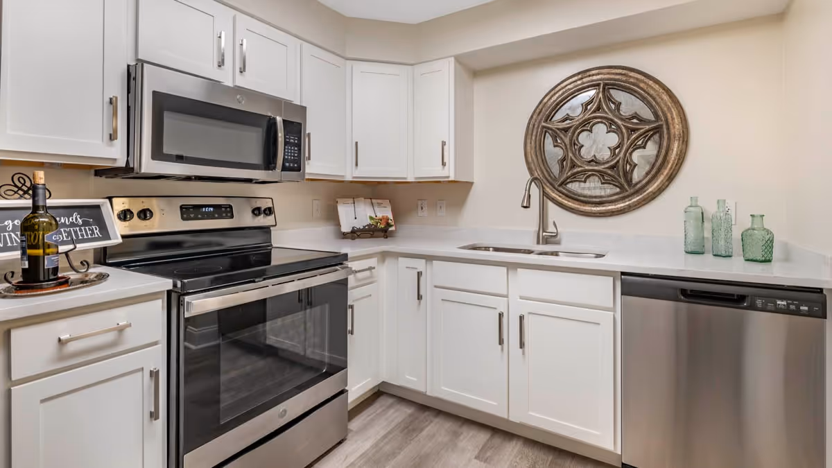 Modern kitchen with white cabinets, stainless steel microwave, oven, and dishwasher. The countertop has a wine bottle, a decorative sign, a cookbook stand, and three green glass bottles. A round decorative mirror hangs on the wall above the sink.