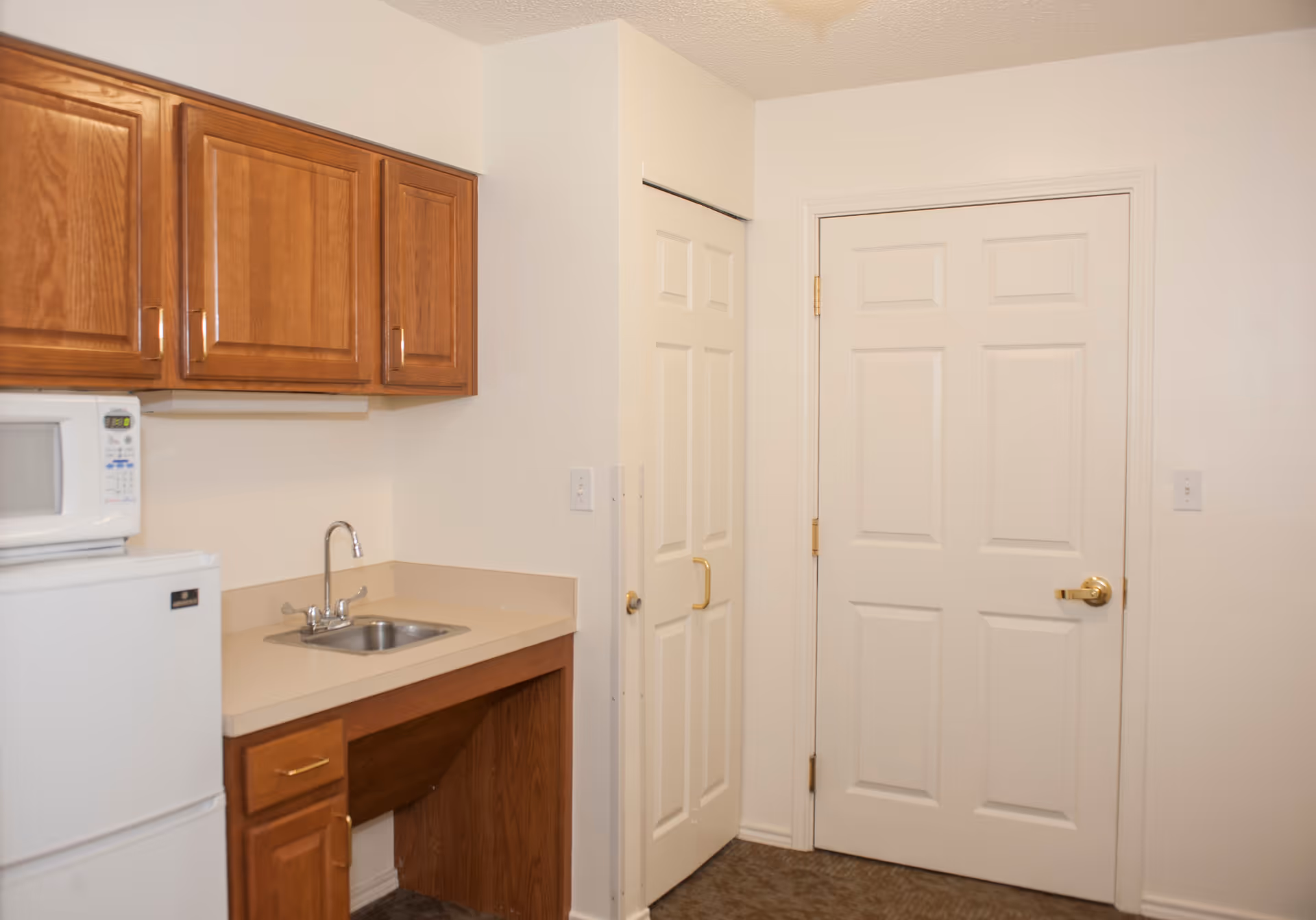 Small kitchenette with wooden cabinets, a sink, a microwave over a mini-fridge, and two white doors.