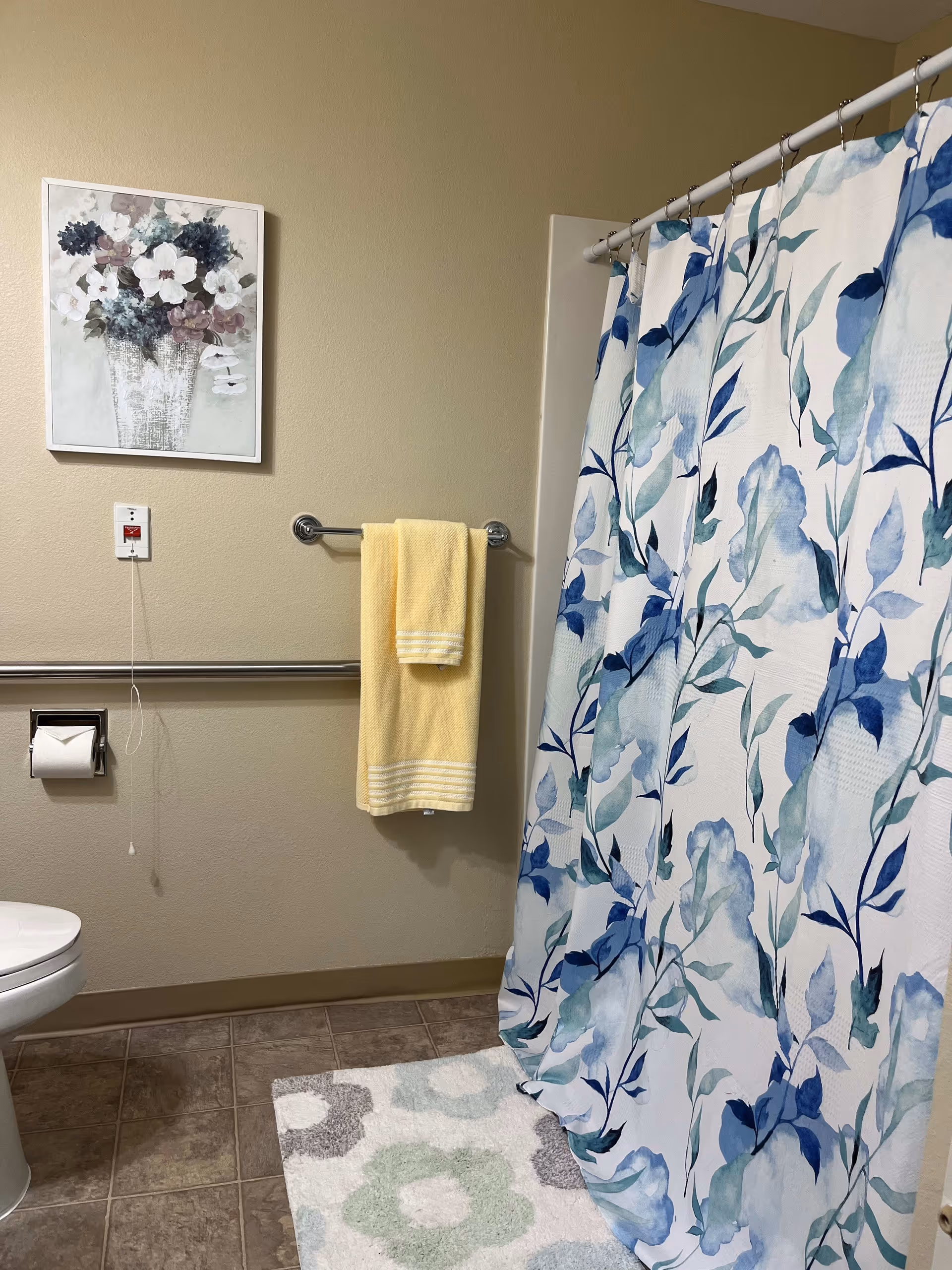Bathroom with a toilet, a floral shower curtain in shades of blue and green, a yellow towel hanging on a towel rack, a floral painting on the wall, a pull cord emergency switch, and a patterned bath mat on the tiled floor.