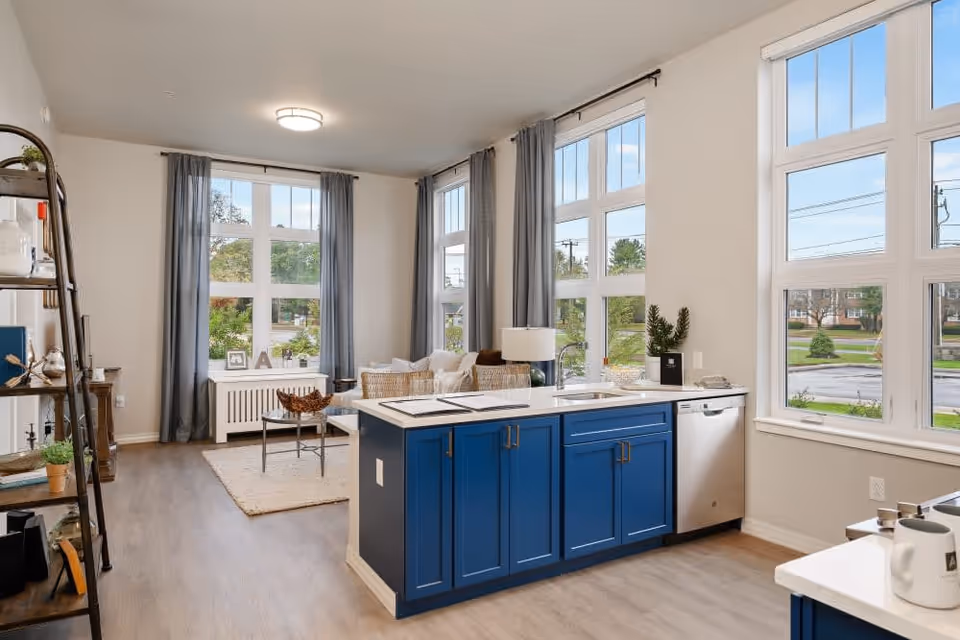 Bright and modern open-concept living space with large windows, blue kitchen island with sink and dishwasher, light wood flooring, and a cozy seating area with a sofa and coffee table near the windows.