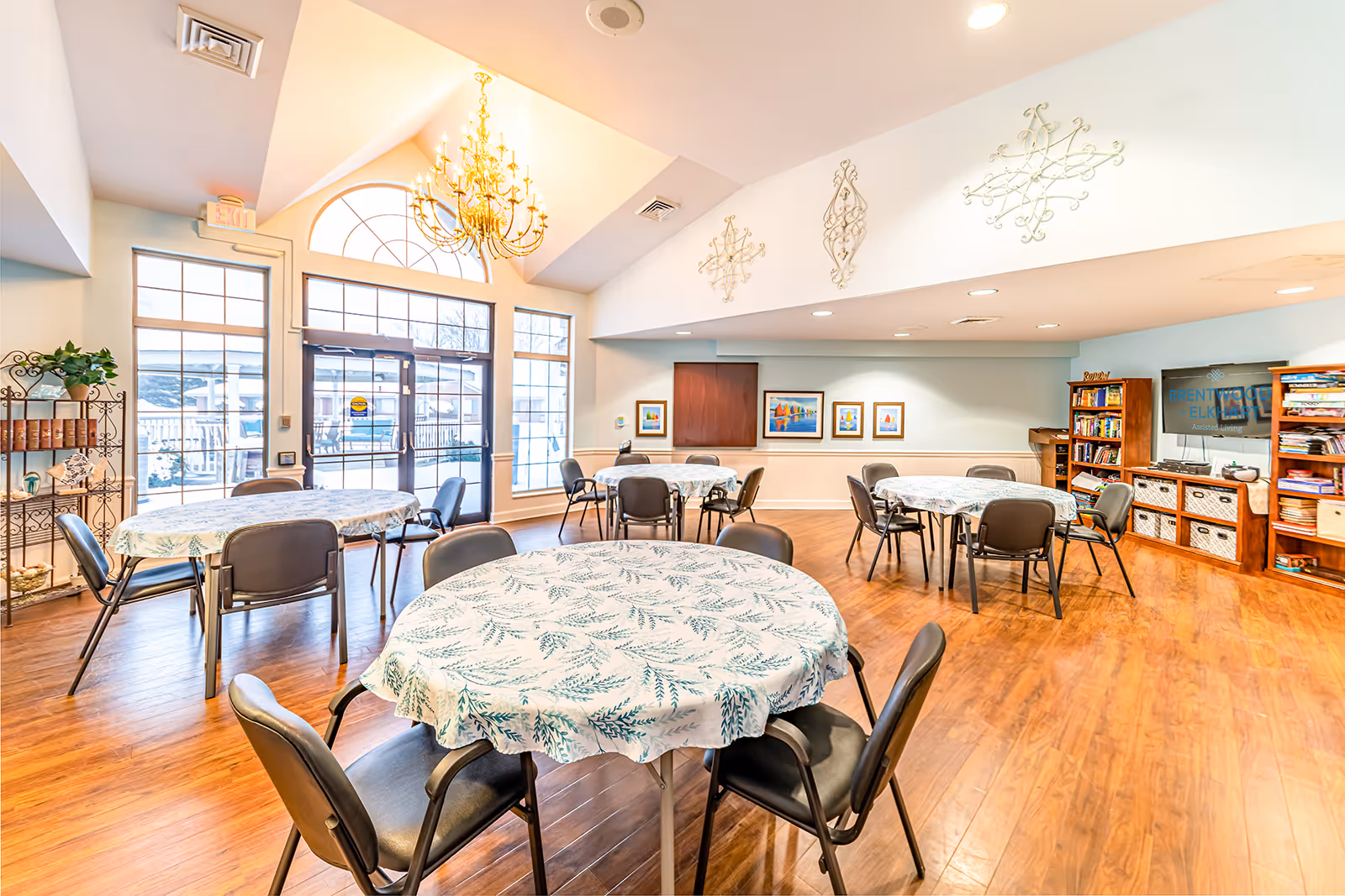 A bright and spacious common area with round tables covered in white tablecloths with green leaf patterns, surrounded by black chairs. The room has large windows and a glass door letting in natural light, a chandelier hanging from a vaulted ceiling, wooden flooring, wall art, and bookshelves filled with books and games. A TV screen displays the text 'Brentwood at Elkhart Assisted Living.'