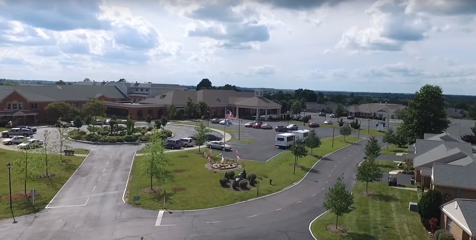 Aerial view of Wesley Village Senior Living facility showing multiple buildings, a circular driveway with landscaped greenery and flags, parked cars, and a bus under a partly cloudy sky.