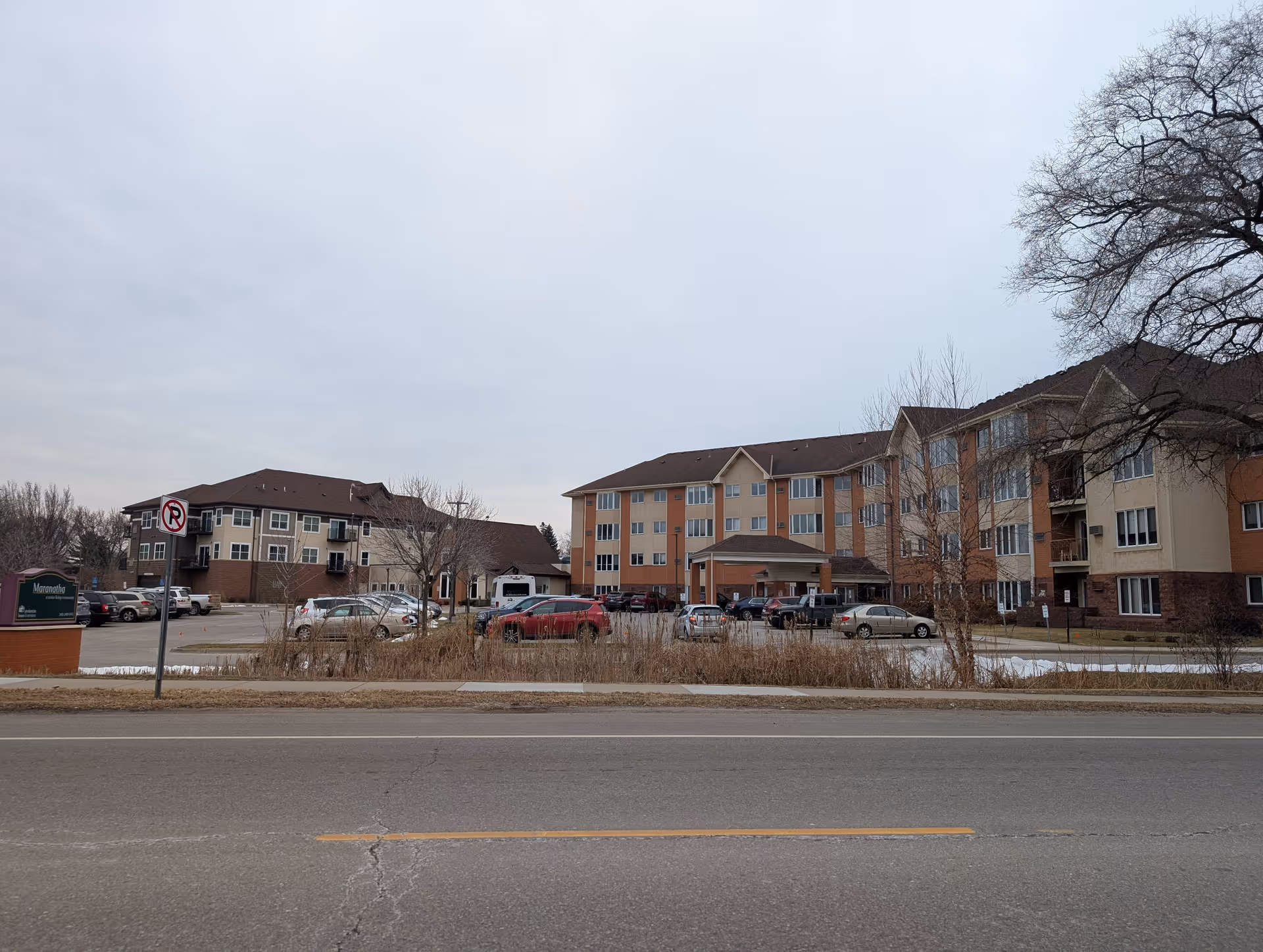 Front exterior of a multi-story senior living building with a parking lot and cars under an overcast sky.