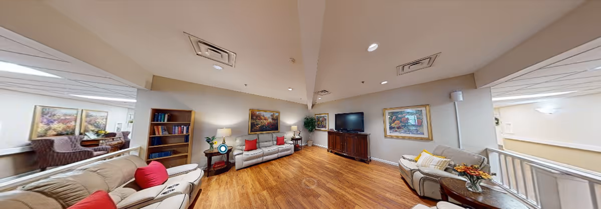 A spacious living room area in an assisted living community featuring beige leather sofas with red and yellow cushions, wooden flooring, a wooden TV cabinet with a flat-screen TV, a bookshelf with books, side tables with lamps, framed artwork on the walls, and a small table with a flower arrangement. The room is well-lit with ceiling lights and natural light from adjacent areas.