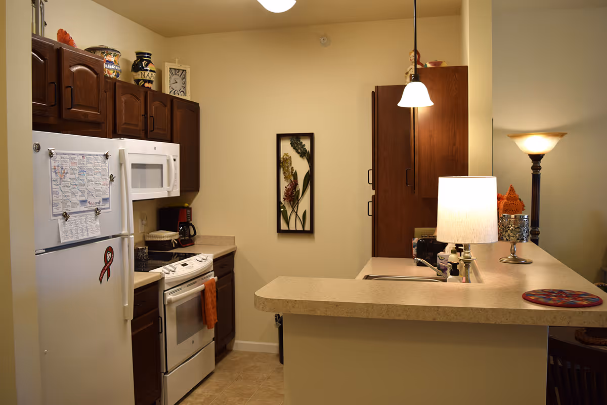 A compact kitchen area with dark wooden cabinets, a white refrigerator with papers and a ribbon magnet on it, a white microwave above a white stove with an orange towel hanging from the handle. The kitchen has beige countertops and a small bar area with a lamp and decorative items. There is a framed floral artwork on the wall and a hanging pendant light above the bar.