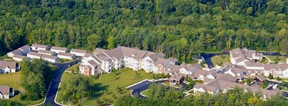 Aerial view of River Terrace Retirement Community surrounded by dense green trees. The facility consists of multiple connected buildings with brown roofs and white walls, arranged in a curved layout with parking areas and driveways.