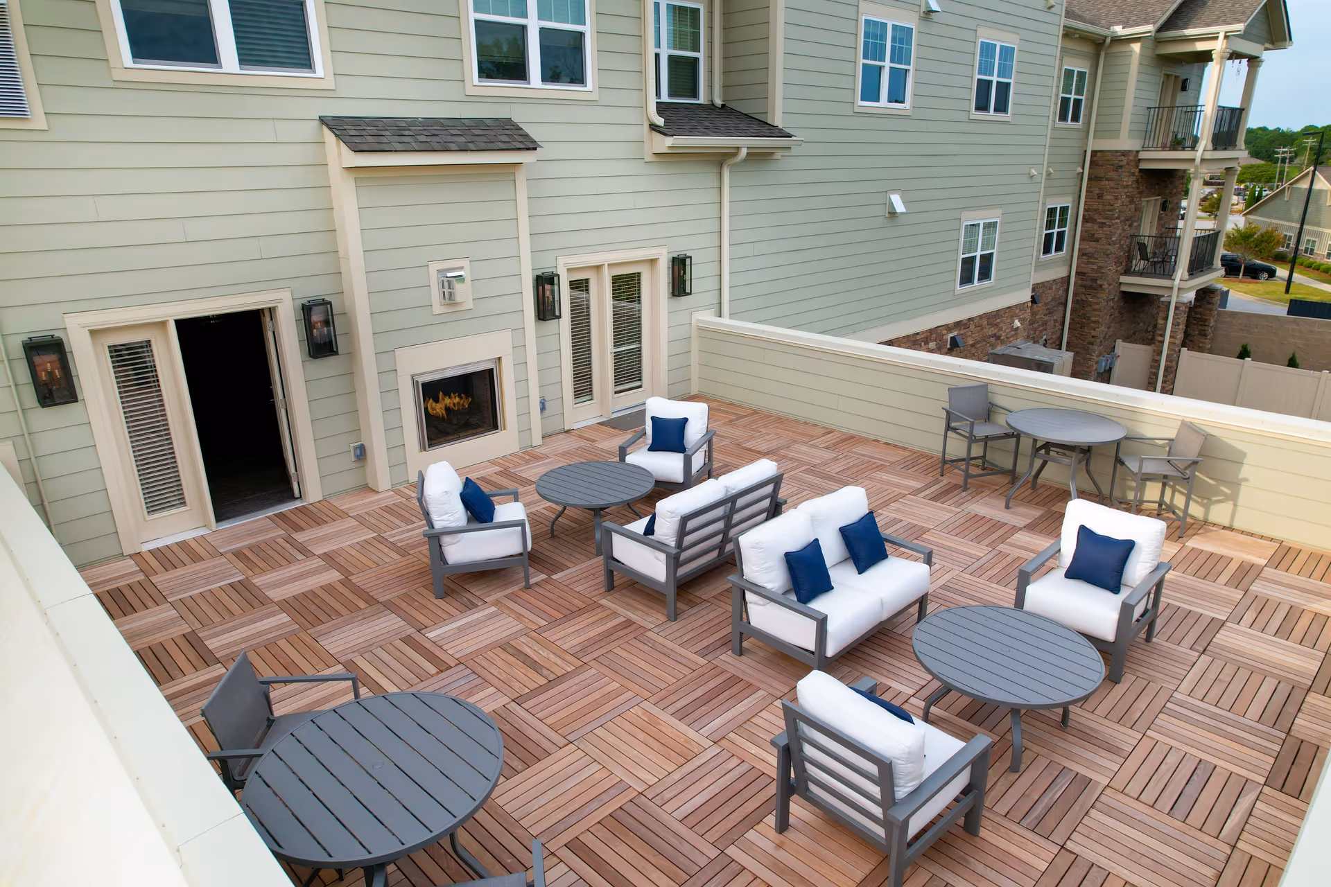 Rooftop patio with wooden tile flooring, multiple seating areas with white cushioned chairs and blue pillows, round tables, and a fireplace against the building.