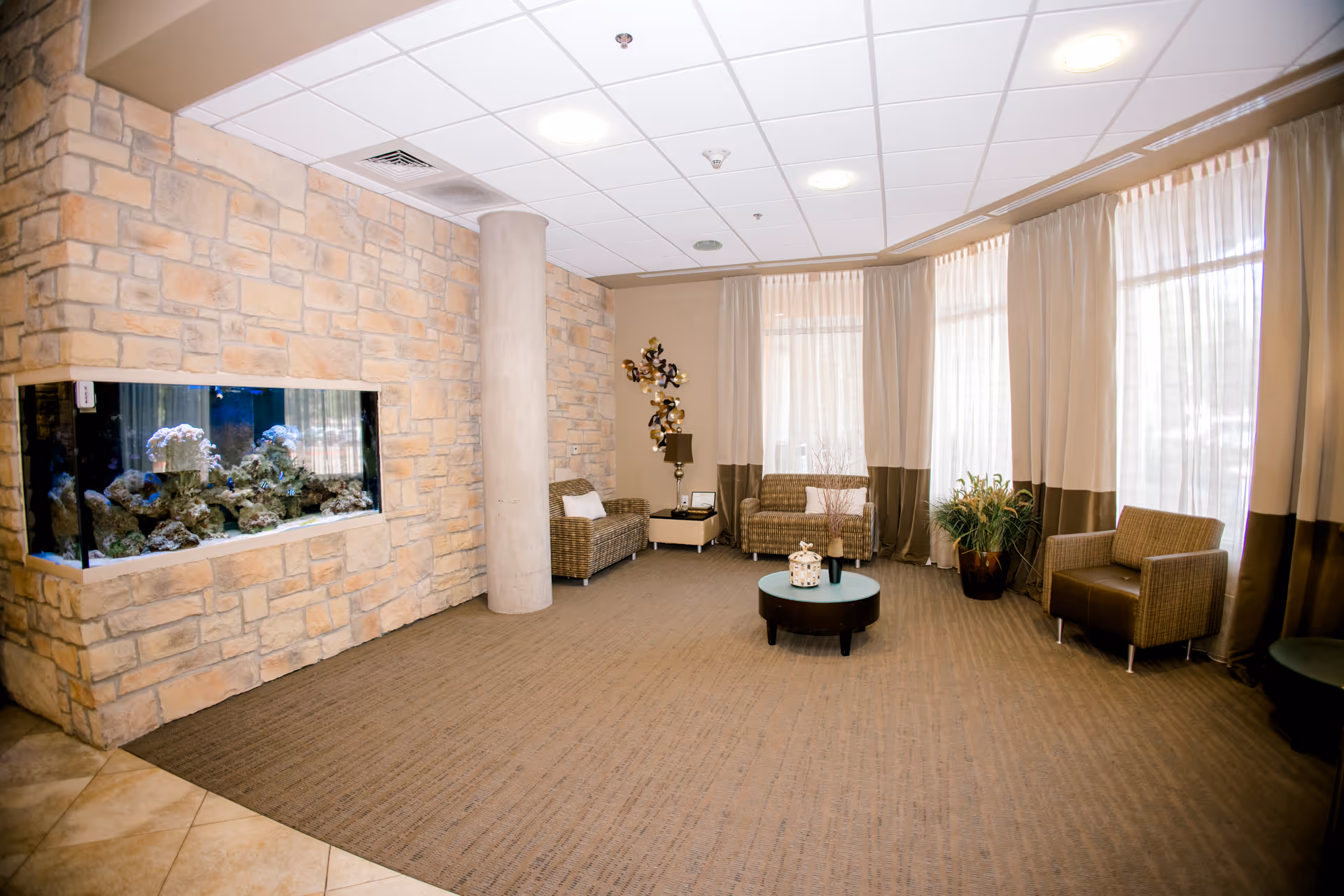A cozy sitting area with wicker chairs and a small round coffee table in the center. The room features a stone wall with a built-in aquarium filled with coral and fish. Large windows with sheer and brown curtains allow natural light to fill the space. There is a decorative wall piece and a potted plant near the windows.