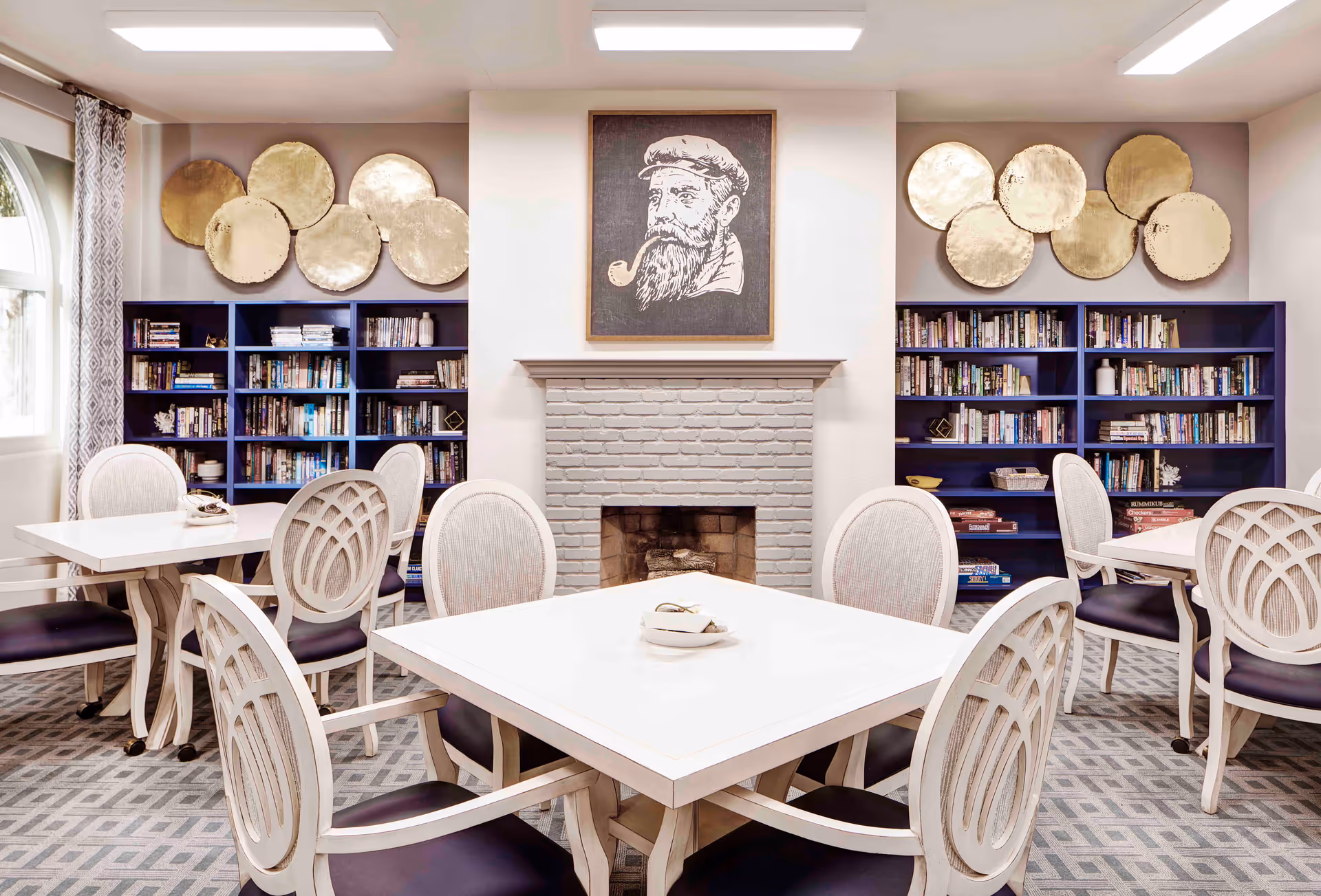 A cozy senior living common area with white tables and chairs arranged around a gray carpeted floor. There is a white brick fireplace in the center with a framed artwork of a bearded man smoking a pipe above it. On either side of the fireplace are blue bookshelves filled with books and decorative items. Above the bookshelves are gold circular wall decorations. A large window with patterned curtains is visible on the left side.
