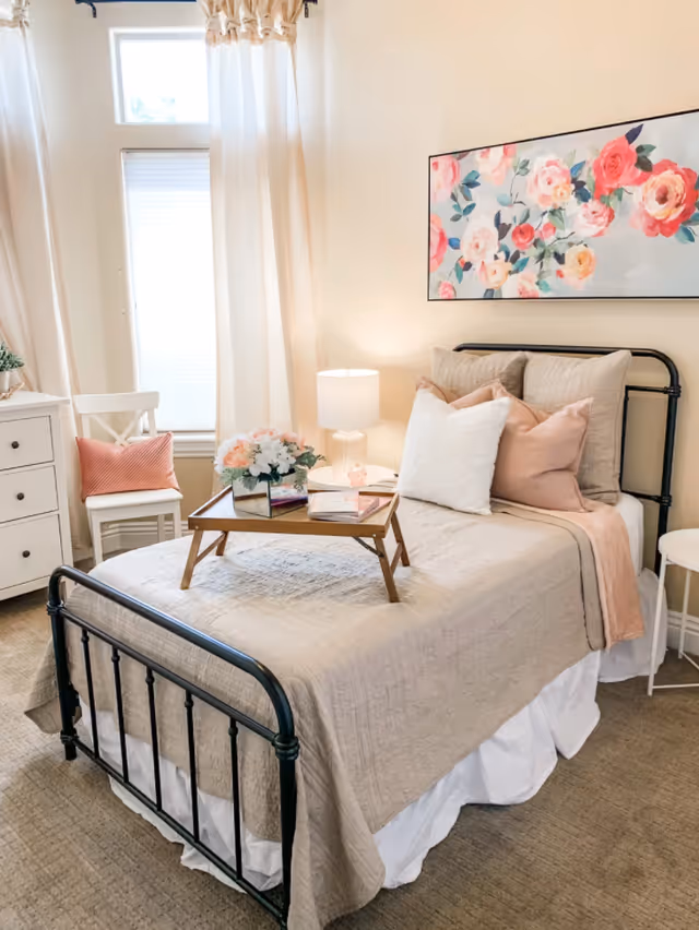 A cozy bedroom with a single black metal bed frame, beige quilted bedspread, and multiple pillows in white and blush tones. A wooden tray with flowers and books sits on the bed. Next to the bed is a small white round side table and a lamp with a white shade. A white chair with a pink cushion is placed near a window with white curtains. A floral painting with pink and red flowers hangs on the wall above the bed.