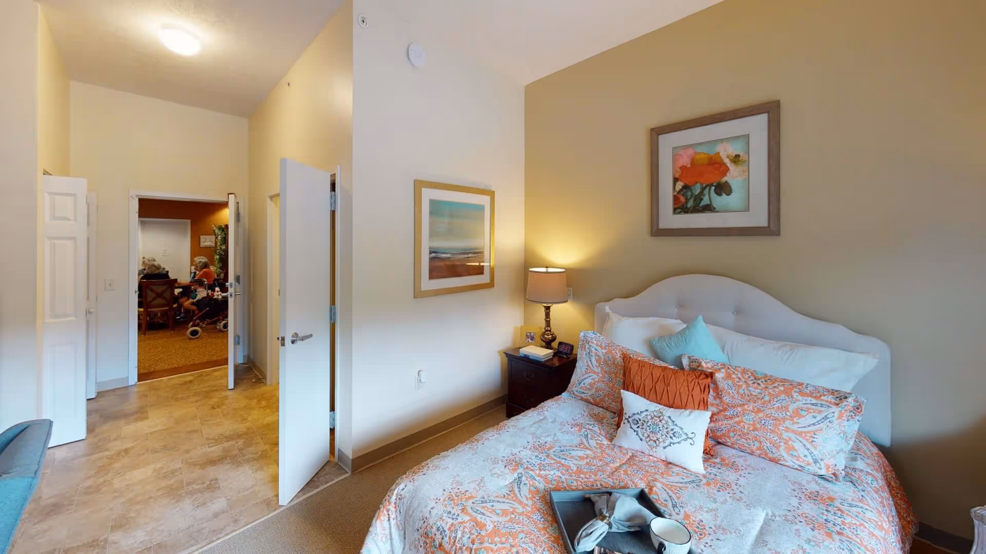 A cozy bedroom in a senior living facility with a bed covered in orange and white patterned bedding, a nightstand with a lamp, and framed artwork on the walls. The door is open, showing a hallway and a room with several elderly people sitting and conversing.