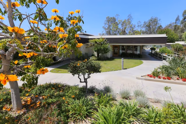 Front entrance of a single-story care facility with a circular driveway, landscaped gardens, and a yellow-flowering tree in the foreground.