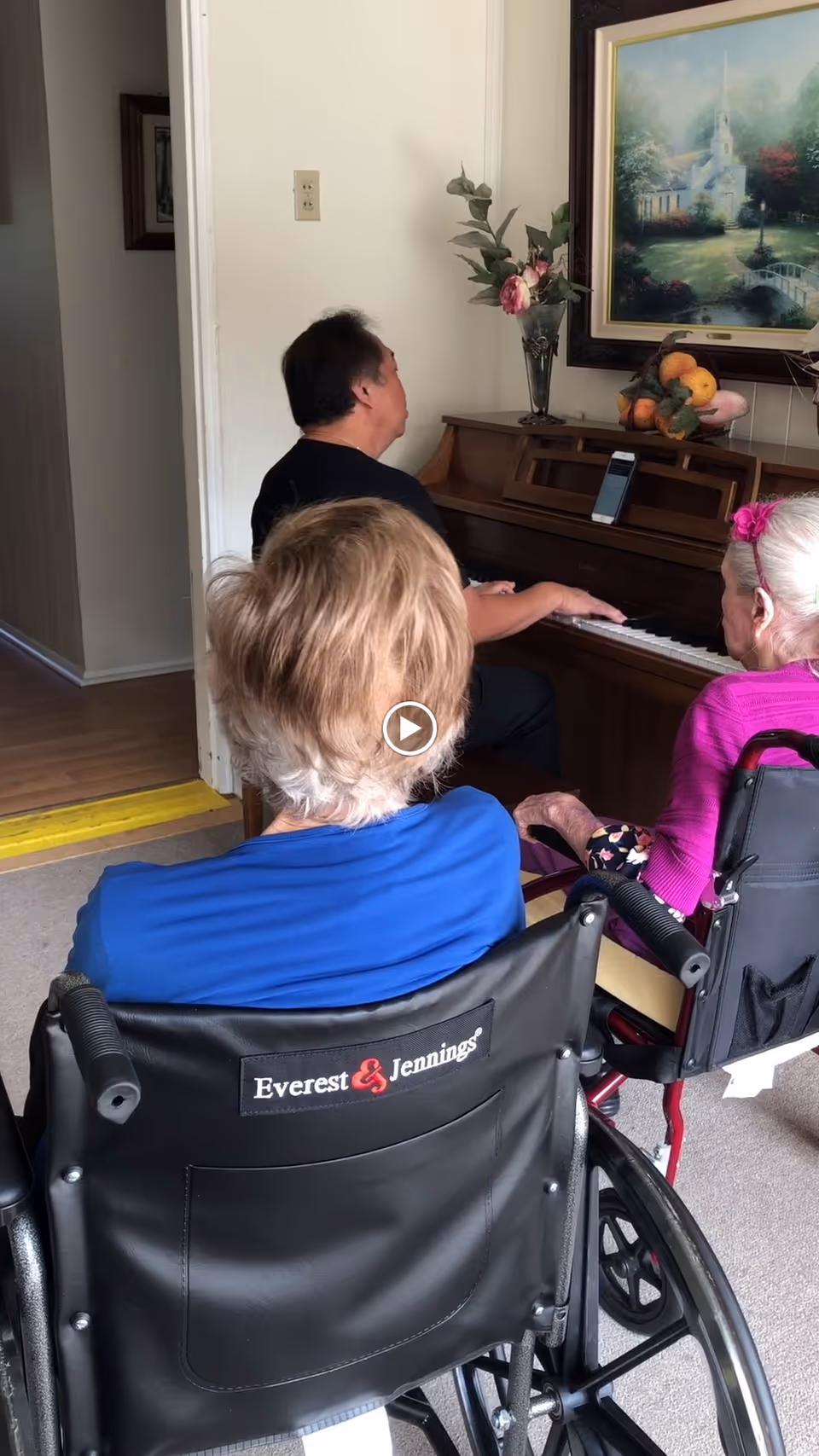 Two elderly women in wheelchairs facing a man playing a piano in a room. The piano has a vase with flowers and a bowl of fruit on top, and a framed painting hangs on the wall above the piano.