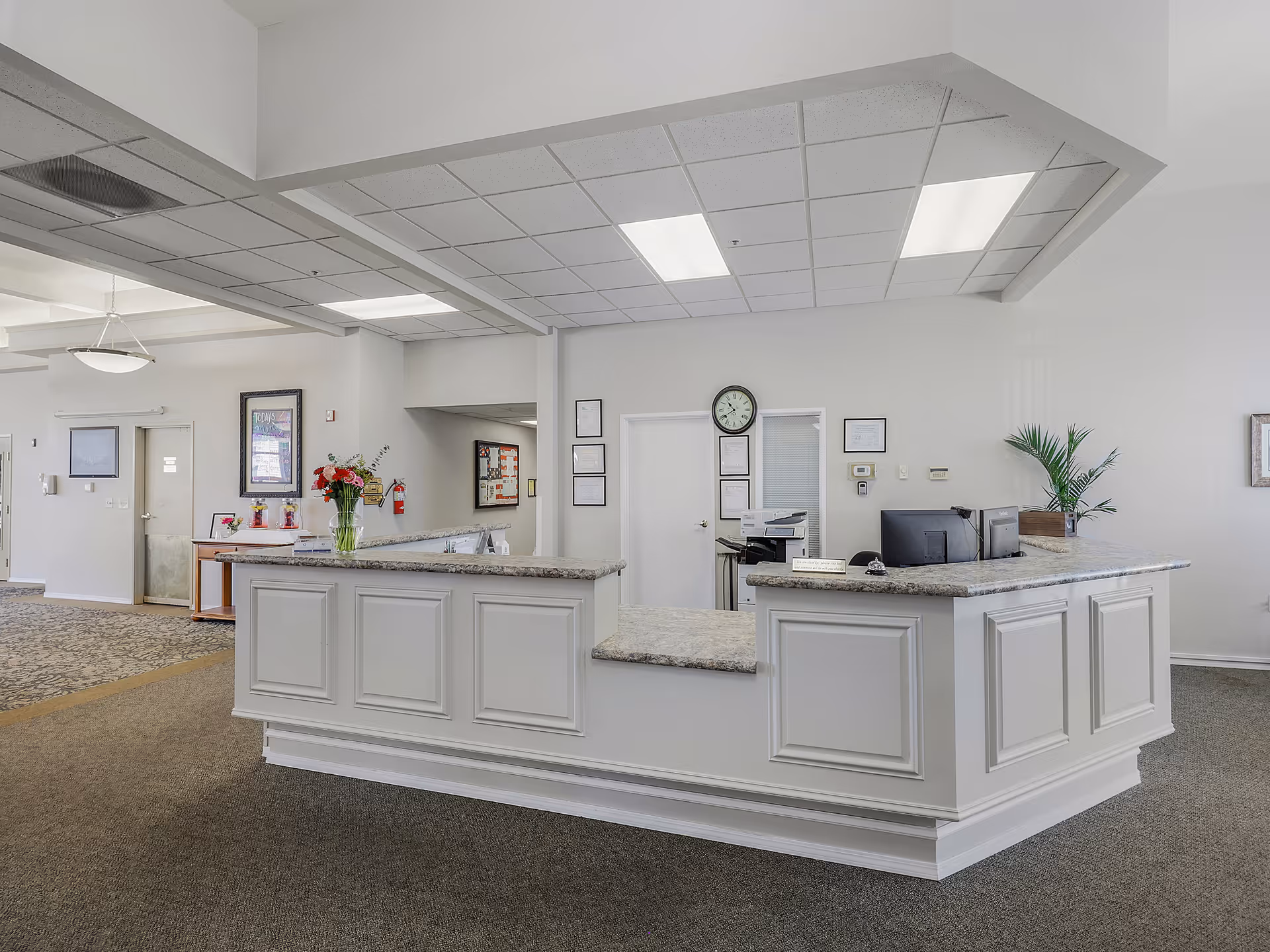 Reception desk in an assisted living facility lobby with granite countertops, a wall clock, and office equipment.