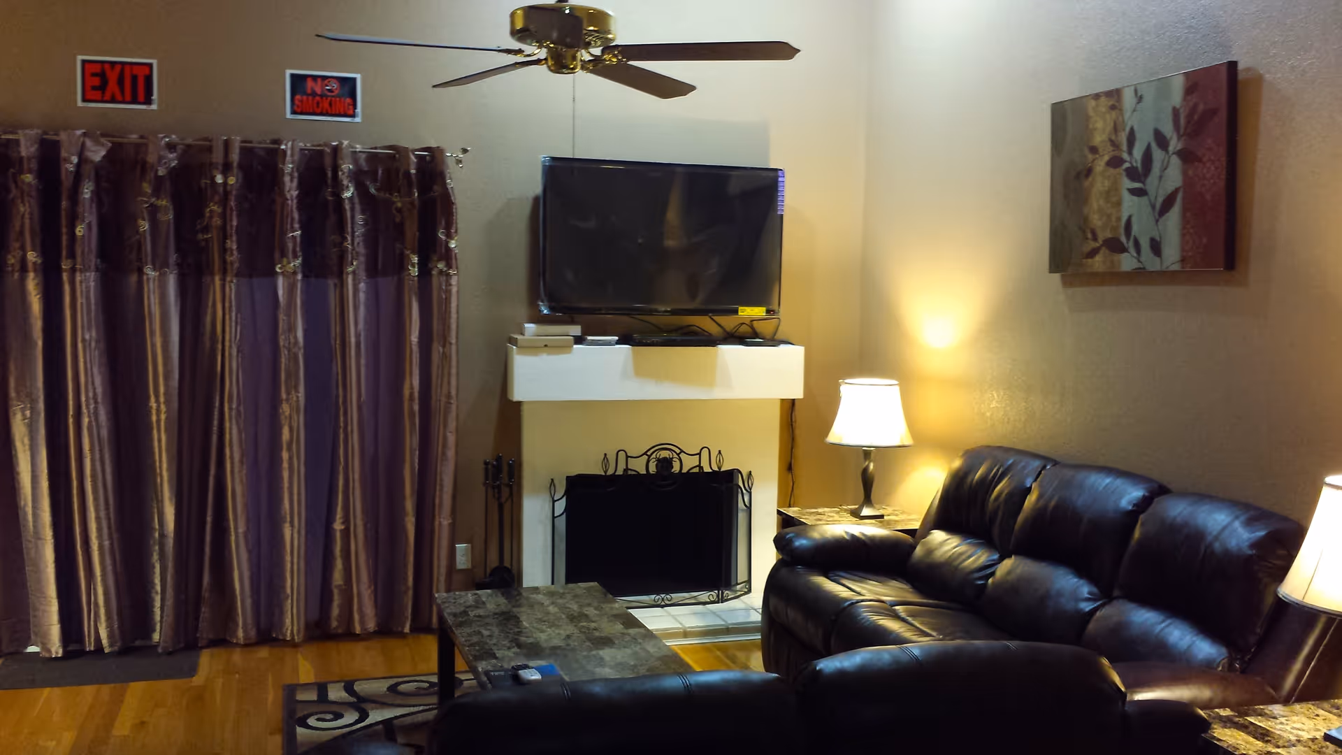 A cozy living room with a dark leather sofa and loveseat, a coffee table, two side tables with lamps, a wall-mounted flat-screen TV above a fireplace with a decorative screen, a ceiling fan, and curtains covering a window or door. There are exit and no smoking signs on the wall above the curtains.