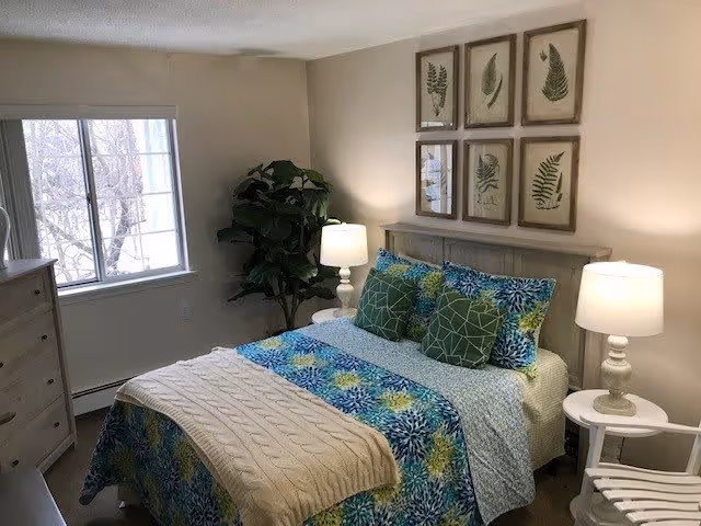 A cozy bedroom with a bed covered in a colorful blue, green, and yellow floral bedspread, accented with green and patterned pillows. There are two white bedside tables, each with a lamp. Above the bed, six framed botanical prints are arranged in two rows. A large window lets in natural light, and a green potted plant is placed in the corner near the window. The room has light-colored walls and wooden flooring.