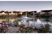 A scenic view of a pond with a water fountain in the center, surrounded by flowering plants and shrubs in the foreground. In the background, there are several single-story residential buildings with pitched roofs under a clear sky.