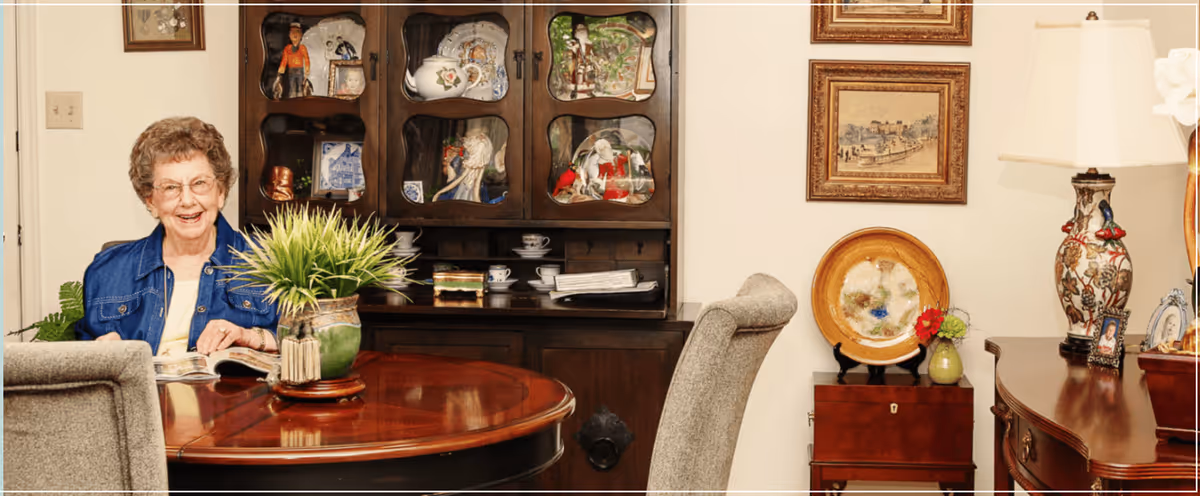An elderly woman with glasses and a blue jacket sitting at a wooden dining table with a green potted plant centerpiece. Behind her is a dark wooden cabinet displaying decorative plates and figurines. The room also features framed artwork on the wall, a table lamp with a floral design, and additional wooden furniture.