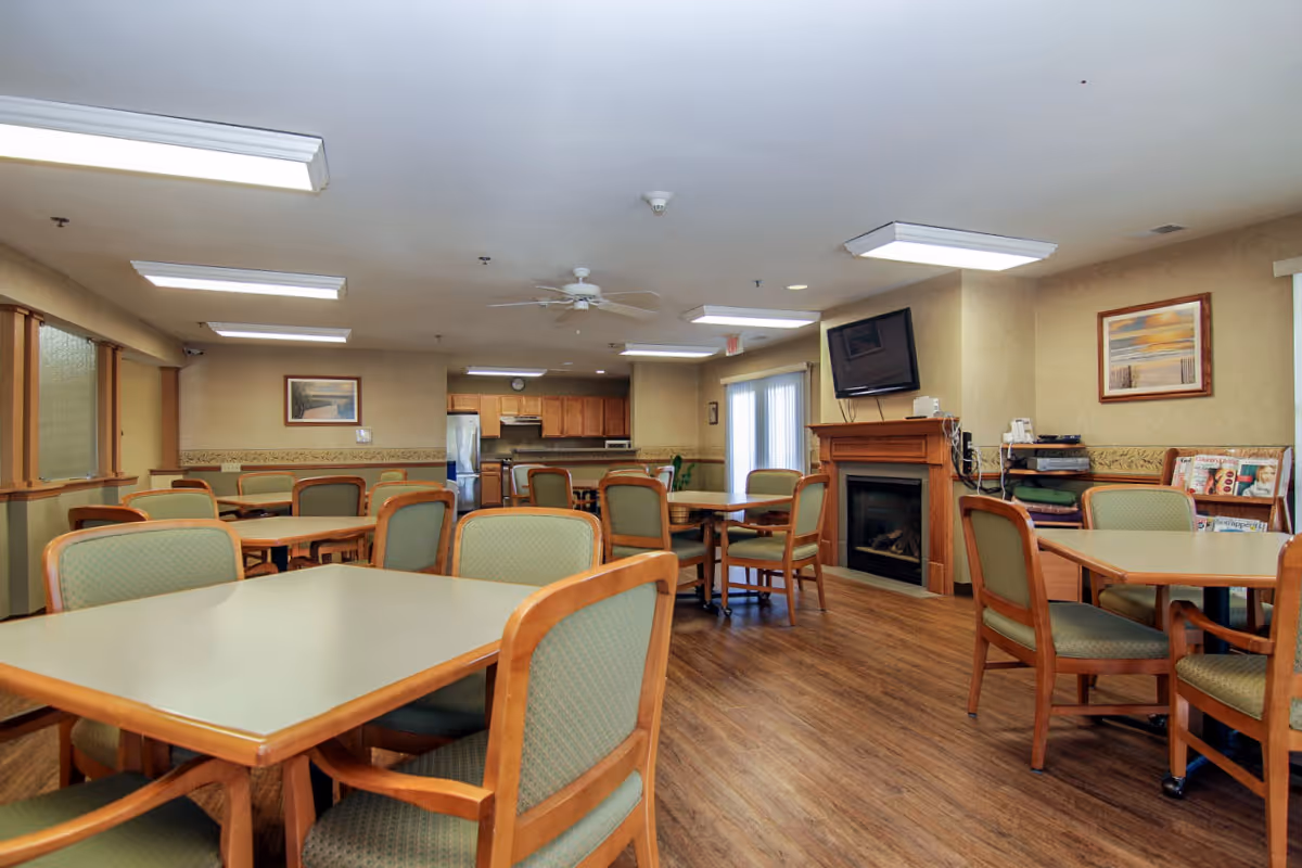 A spacious dining room in Villa Ciera Senior Apartments featuring multiple tables with green cushioned chairs. The room has wood flooring, beige walls, and ceiling lights. There is a fireplace with a mounted TV above it on the right side, and a kitchen area with wooden cabinets and stainless steel appliances in the background. Framed artwork and a magazine rack are also visible.