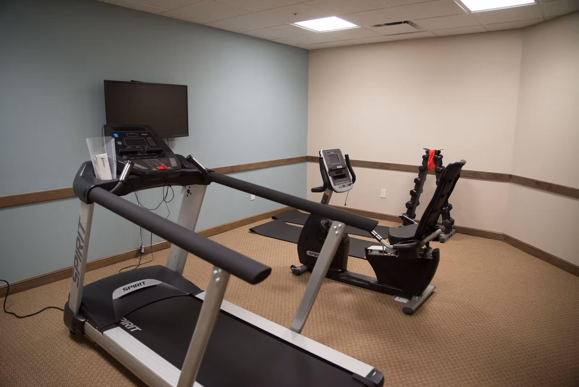 Small fitness room with a treadmill, a recumbent exercise bike, a rack of dumbbells, and two exercise mats on a carpeted floor. The walls are painted in light blue and beige with a wooden chair rail, and a flat screen TV is mounted on the wall above the treadmill.