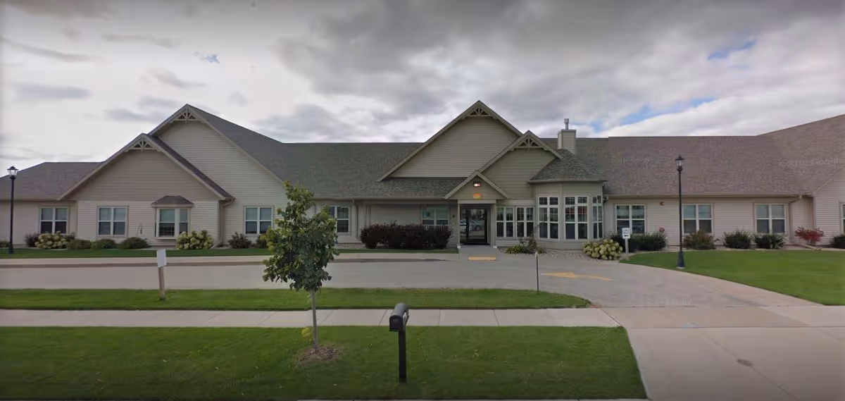 Front exterior of a single-story senior living facility with gabled roofs, central entrance canopy, driveway and manicured lawn.