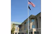 Exterior front of a multi-story beige building with American and POW flags flying on a flagpole against a clear blue sky.