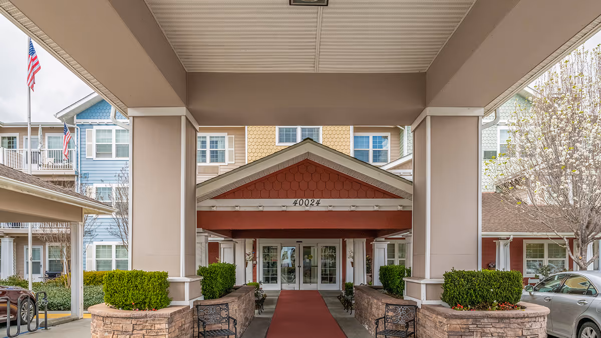 Covered entrance and porte-cochère of a multistory senior living building with a red carpeted walkway and columns leading to glass doors.
