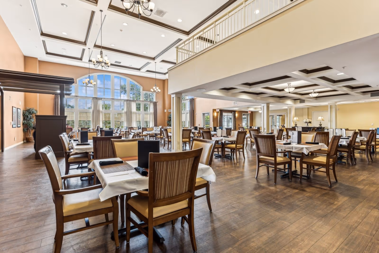 Spacious dining room with multiple wooden tables and chairs arranged neatly, large windows allowing natural light to fill the room, chandeliers hanging from the ceiling, and a warm, inviting atmosphere.