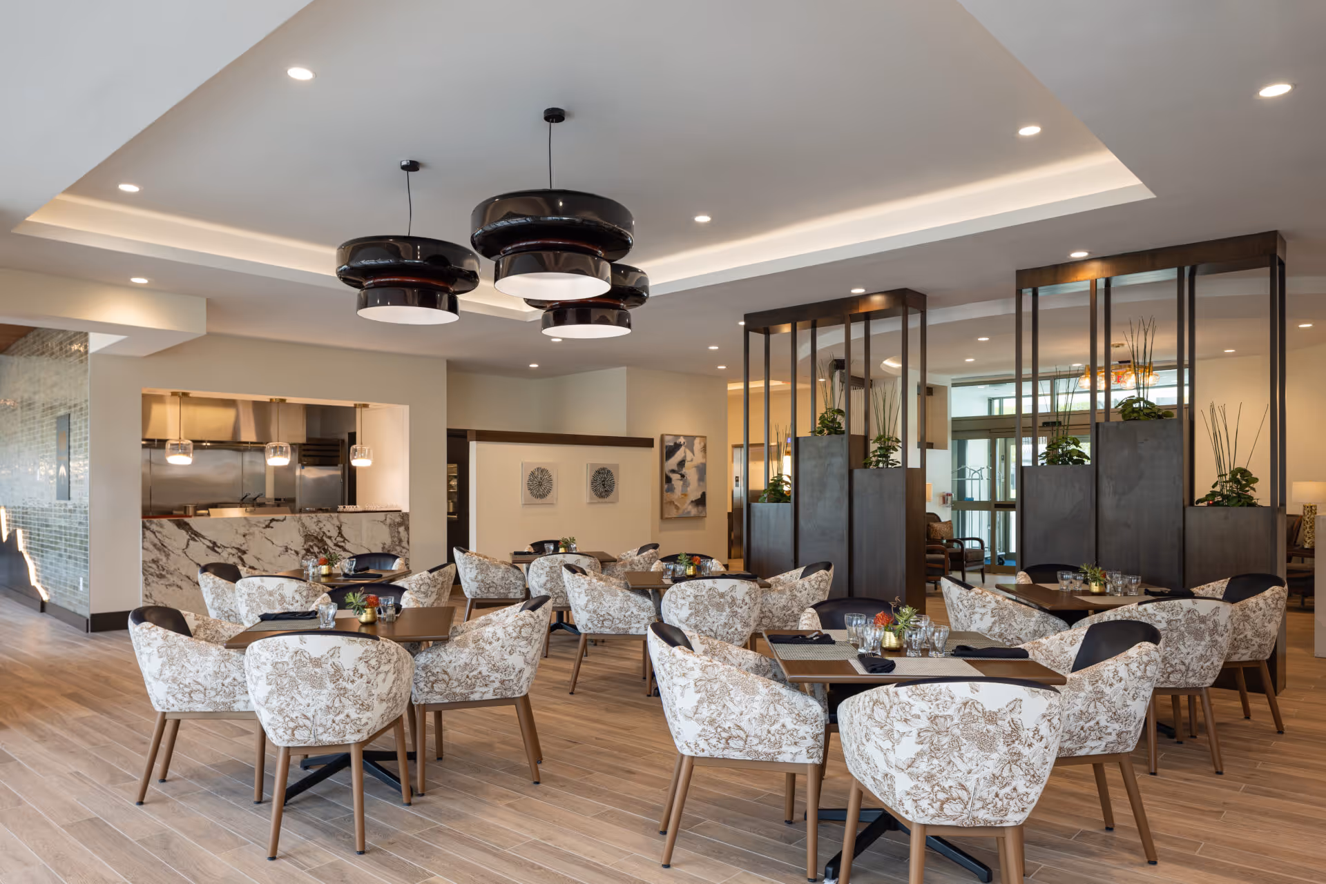 A modern dining area with multiple tables and cushioned chairs featuring floral upholstery. The room has wooden flooring, pendant lights hanging from a recessed ceiling, and a marble counter in the background. Decorative plants are placed on tall wooden dividers, and artwork is displayed on the walls.