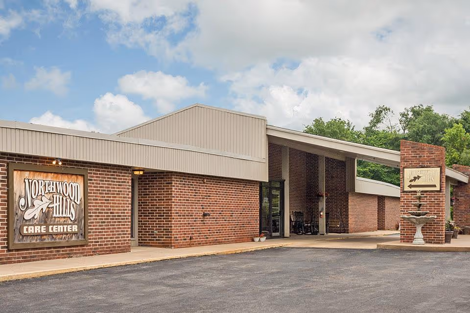 Exterior view of Northwood Hills Care Center, a single-story brick building with a beige roof. The entrance area features a covered walkway with rocking chairs and a decorative fountain near a brick column with the facility's sign. The sky is partly cloudy with some greenery in the background.