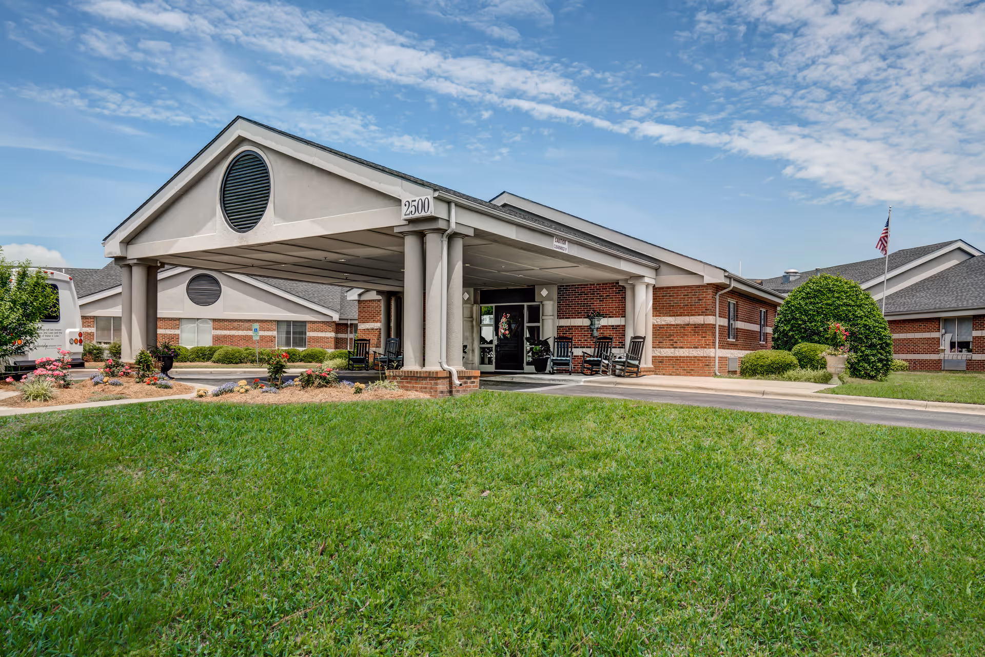 Front entrance of a single-story brick retirement center with a covered porte-cochere, rocking chairs, and a manicured lawn.