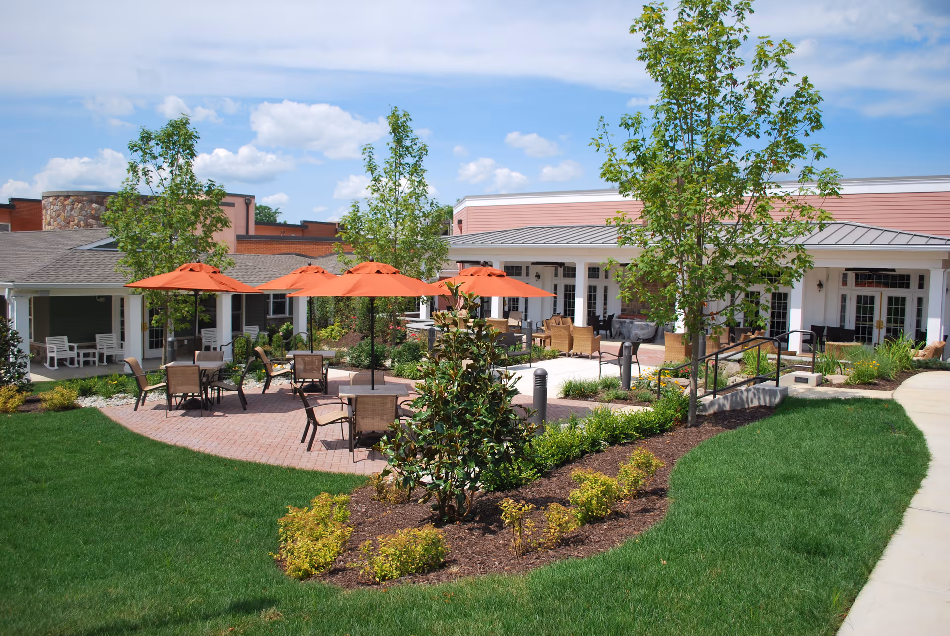 Outdoor patio area at Heath Village Retirement Community with several tables and chairs under orange umbrellas, surrounded by green grass, trees, and landscaped plants. The building with a covered porch and multiple doors is visible in the background under a partly cloudy blue sky.