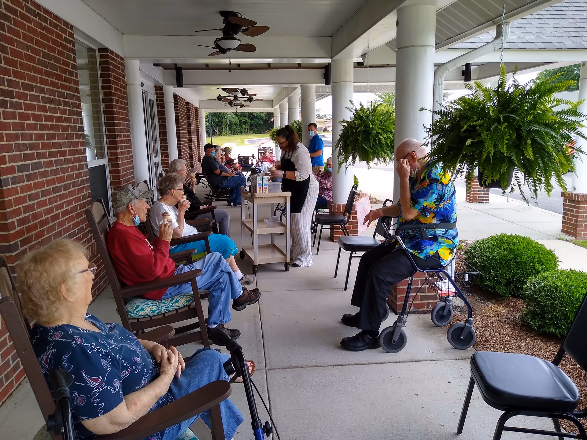 A group of elderly people sitting on rocking chairs along a covered outdoor walkway of a brick building. Some are drinking from cups while a caregiver wearing an apron serves drinks from a cart. There are ceiling fans above and hanging green plants on the right side near white columns. One elderly man is sitting with a walker beside him.