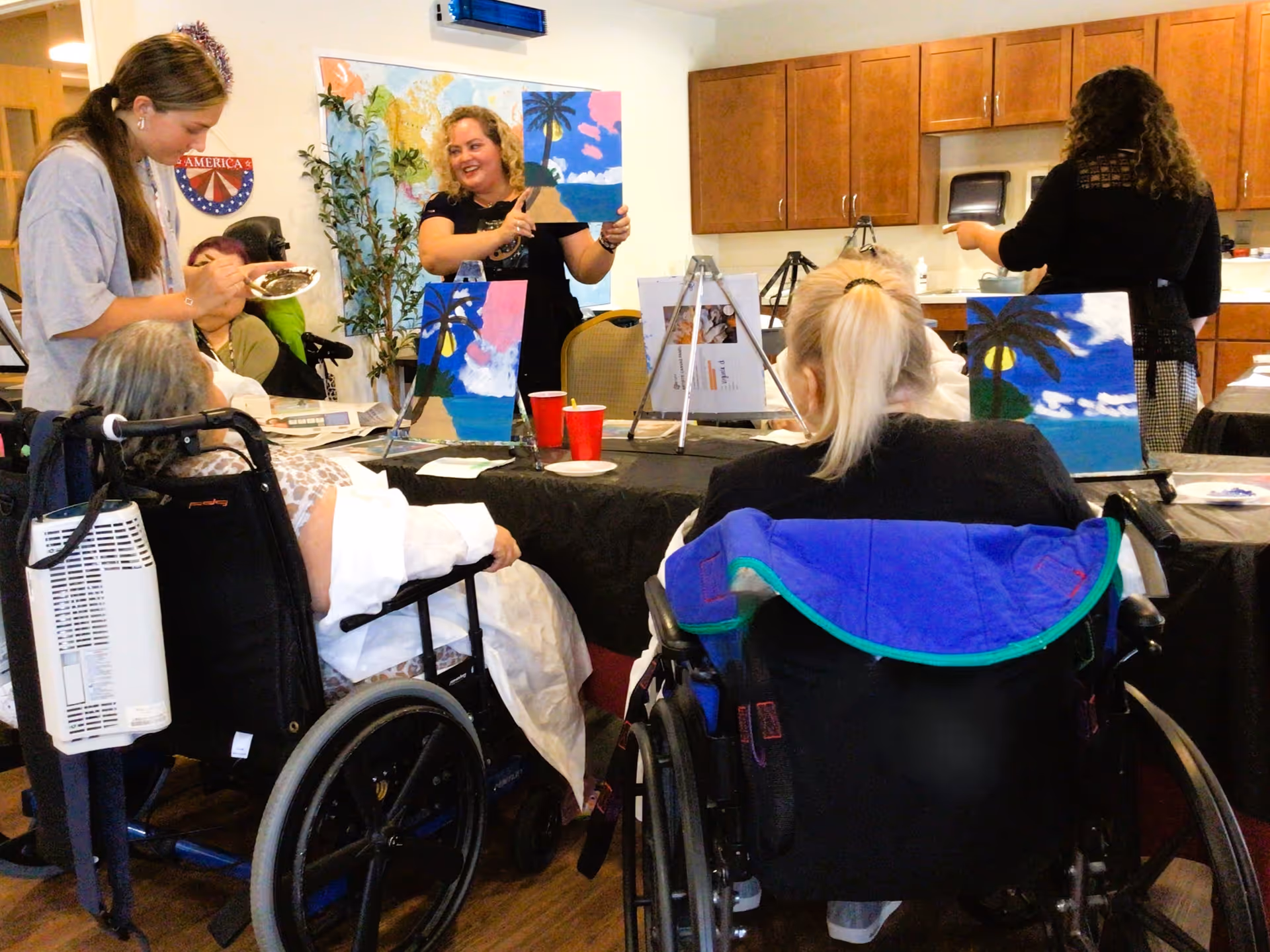 A group of elderly individuals in wheelchairs participating in a painting activity at a table inside a room with wooden cabinets. Two women are standing, one showing a painting of a beach scene with palm trees, while the others are seated and working on similar paintings.