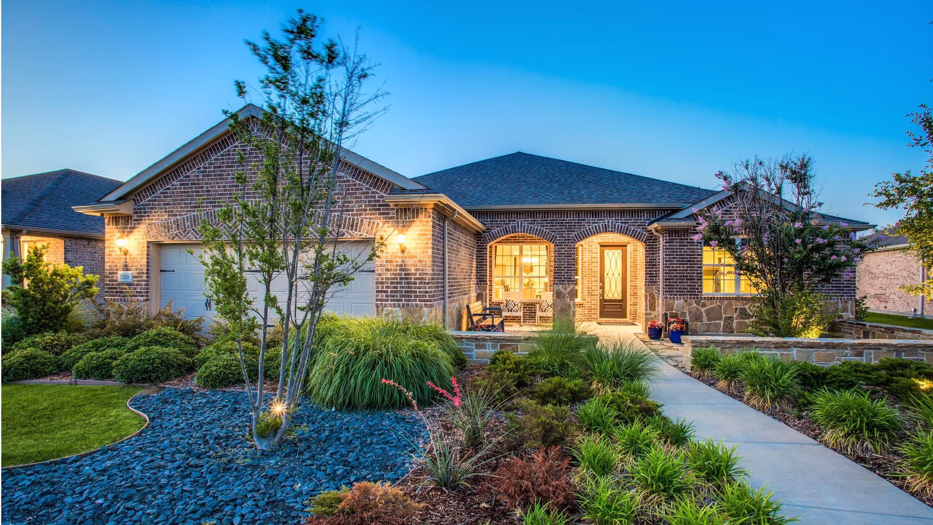 Front exterior view of a single-story brick house with a two-car garage, well-maintained landscaping including bushes, trees, and a stone-lined garden bed, and a concrete walkway leading to a front porch with seating. The house is illuminated with warm lights during dusk.