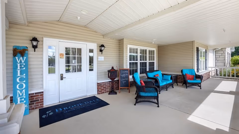 Covered outdoor porch area at Brookdale Goodlettsville featuring a white double door entrance with glass panels, a blue welcome sign, a black doormat with the Brookdale logo, black wicker chairs and loveseat with bright blue cushions and red pillows, a small chalkboard sign, and beige siding with brick accents.