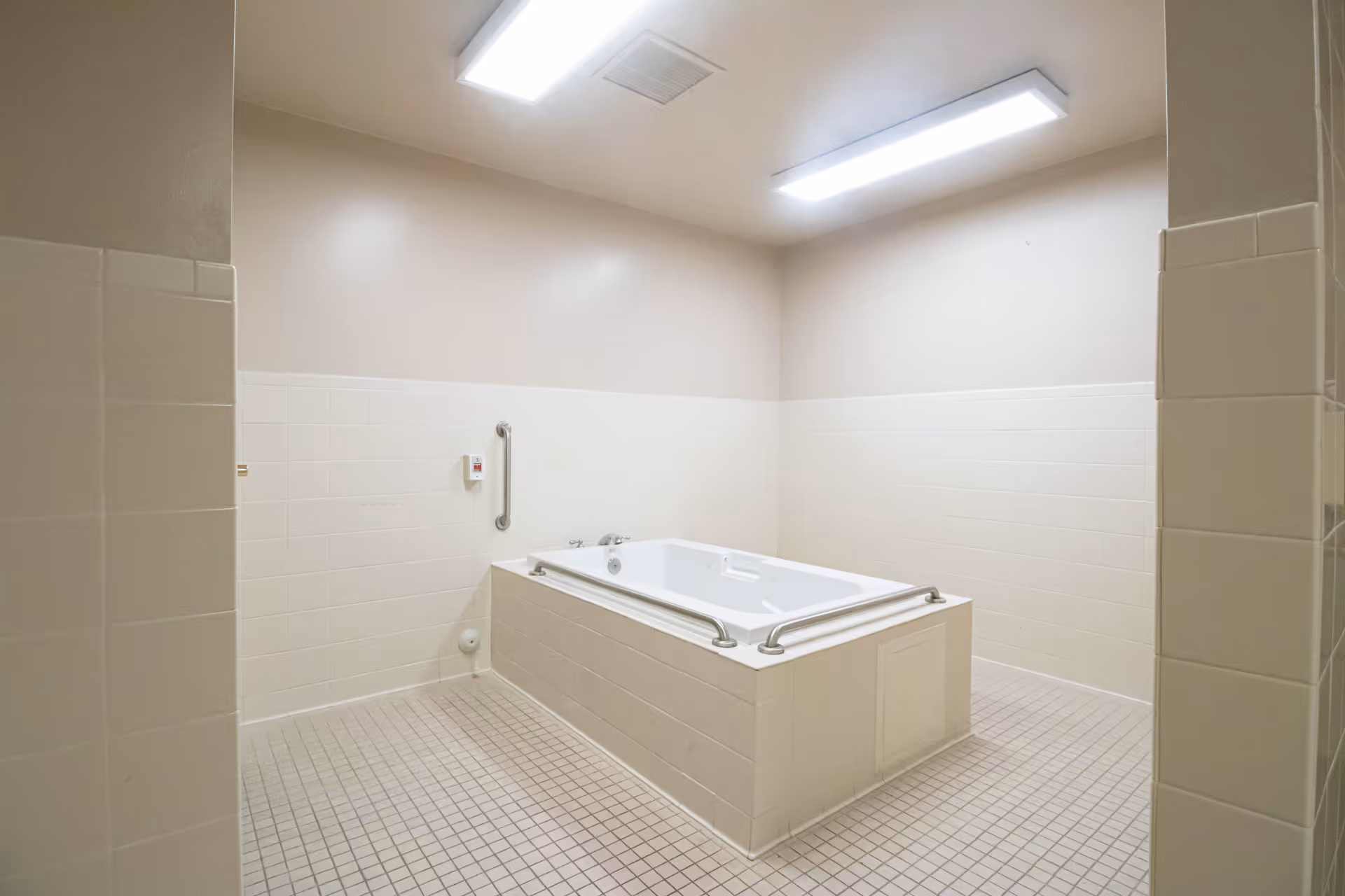 A clean, spacious bathing room with a large rectangular bathtub in the center. The walls are tiled halfway up in white, and the floor is covered with small square tiles. There are grab bars on the bathtub and a control panel on the wall, with bright overhead fluorescent lighting.