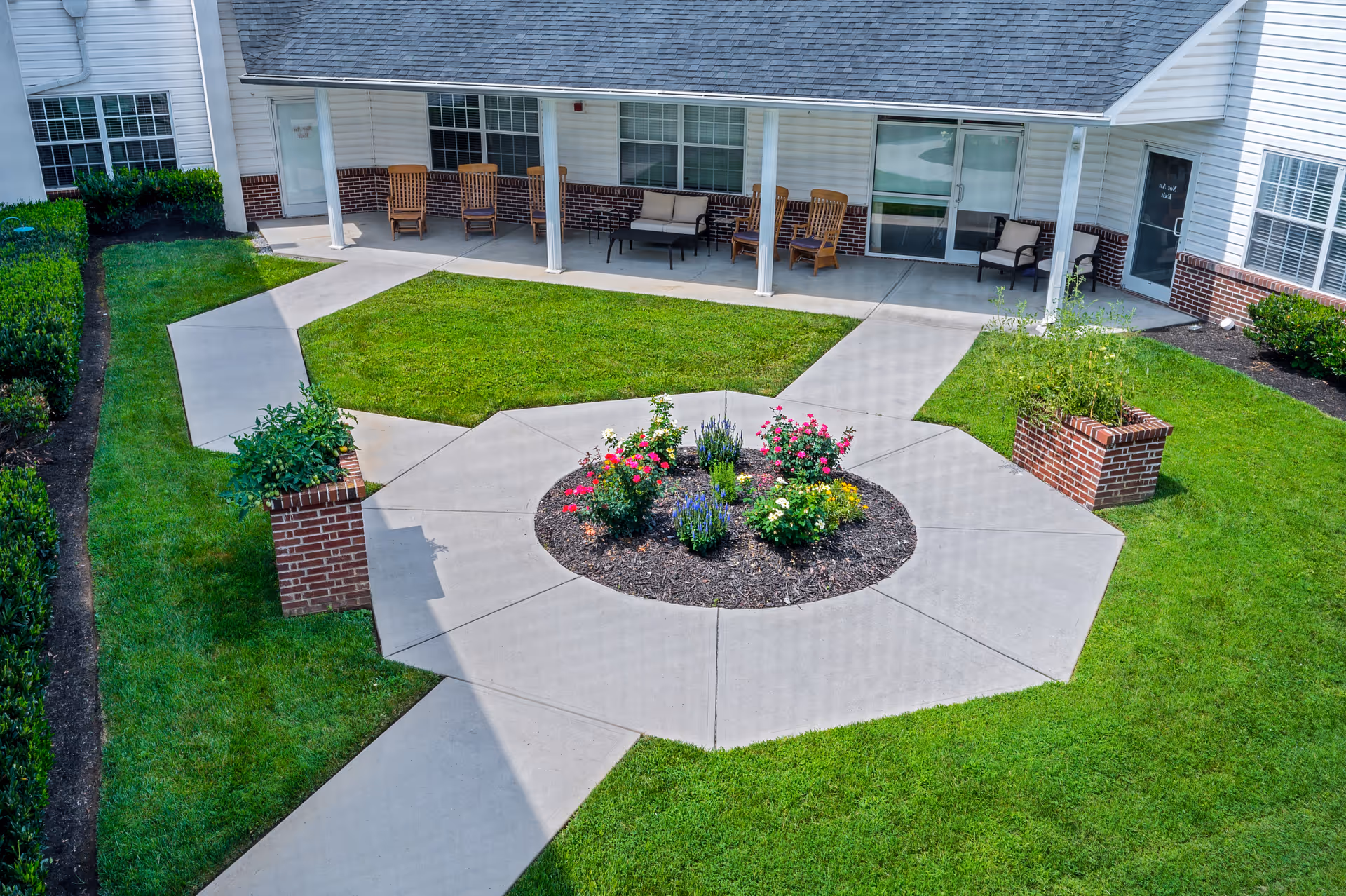 Outdoor courtyard area with a central circular flower bed surrounded by a paved walkway. The courtyard is bordered by green grass and has brick planters with plants. There is a covered patio with several wooden rocking chairs and cushioned seating along the building wall.