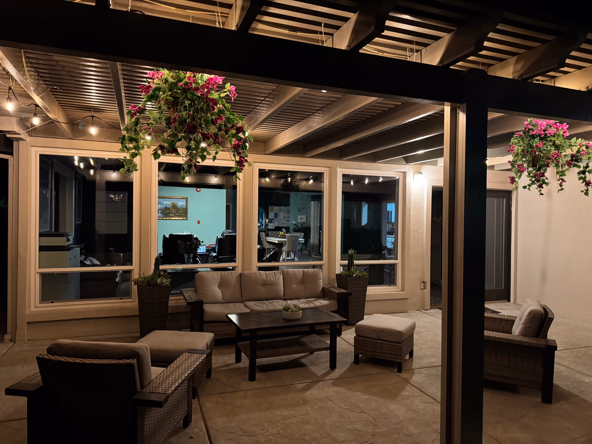 Covered outdoor patio at night with hanging plants, wicker seating, a coffee table, and string lights in front of large windows.