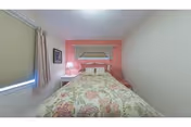 Small bedroom with a bed against a pink accent wall, floral bedspread, window with blinds, and a bedside lamp.